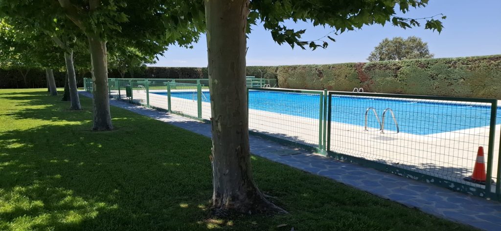 Vaso grande de la piscina municipal visto desde el césped, a la sombra de los árboles